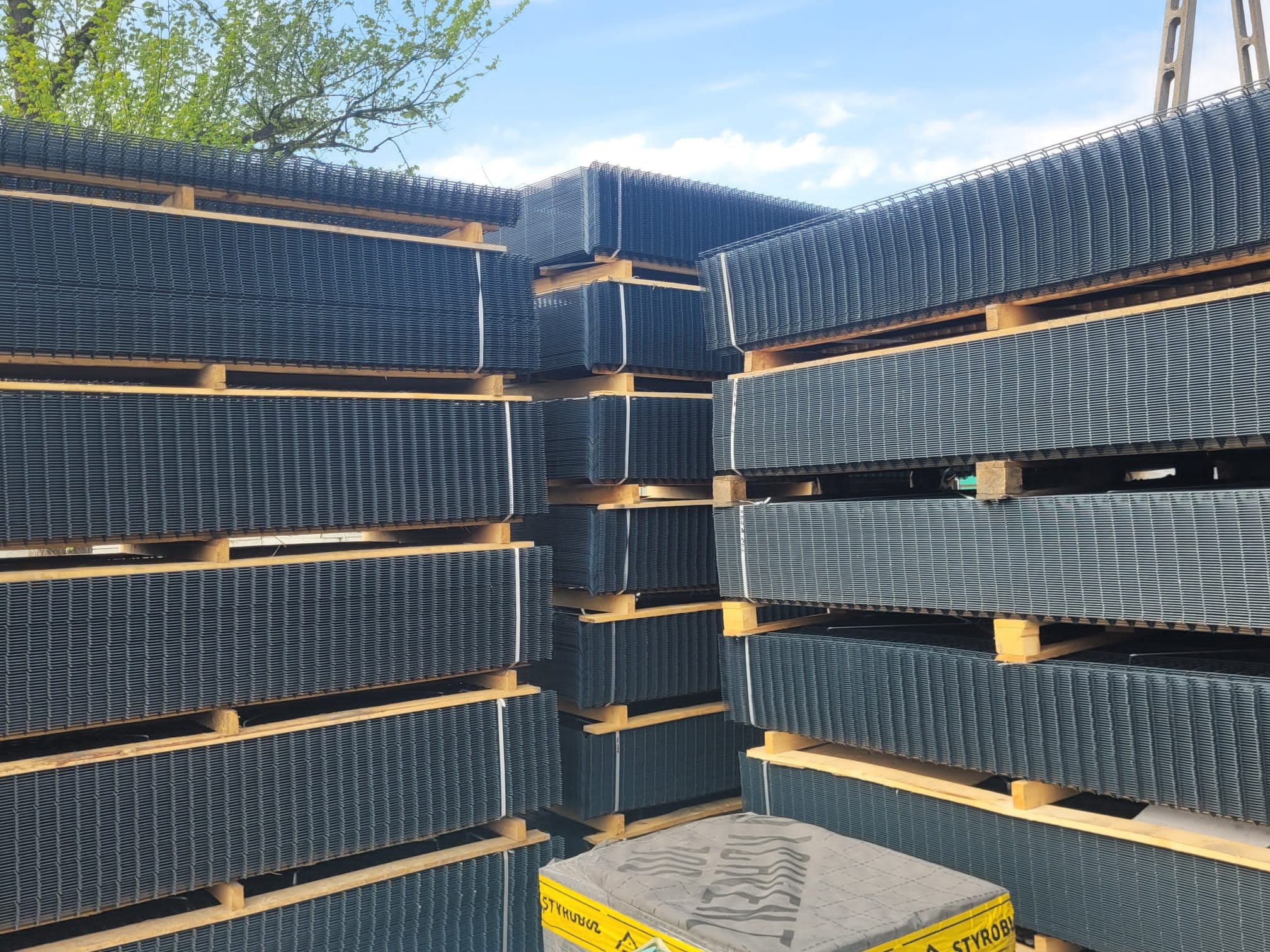 Stos paneli ogrodzeniowych zgrzewanych, ułożonych na drewnianych paletach, zabezpieczonych folią, widok z dołu na tle błękitnego nieba i zieleni drzew.