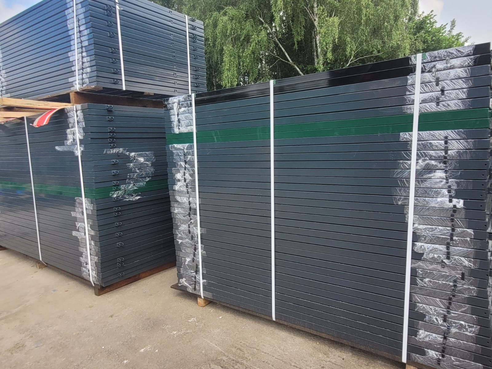 Stosy ciemnoszarych, metalowych paneli ogrodzeniowych zabezpieczonych folią i taśmą na drewnianych paletach, widoczne drzewa w tle.