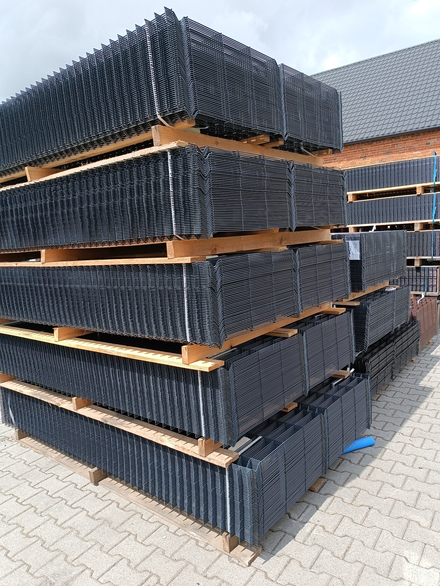Stosy czarnych, metalowych paneli ogrodzeniowych ułożonych na drewnianych paletach na zewnątrz budynku z cegły i dachem z dachówki.