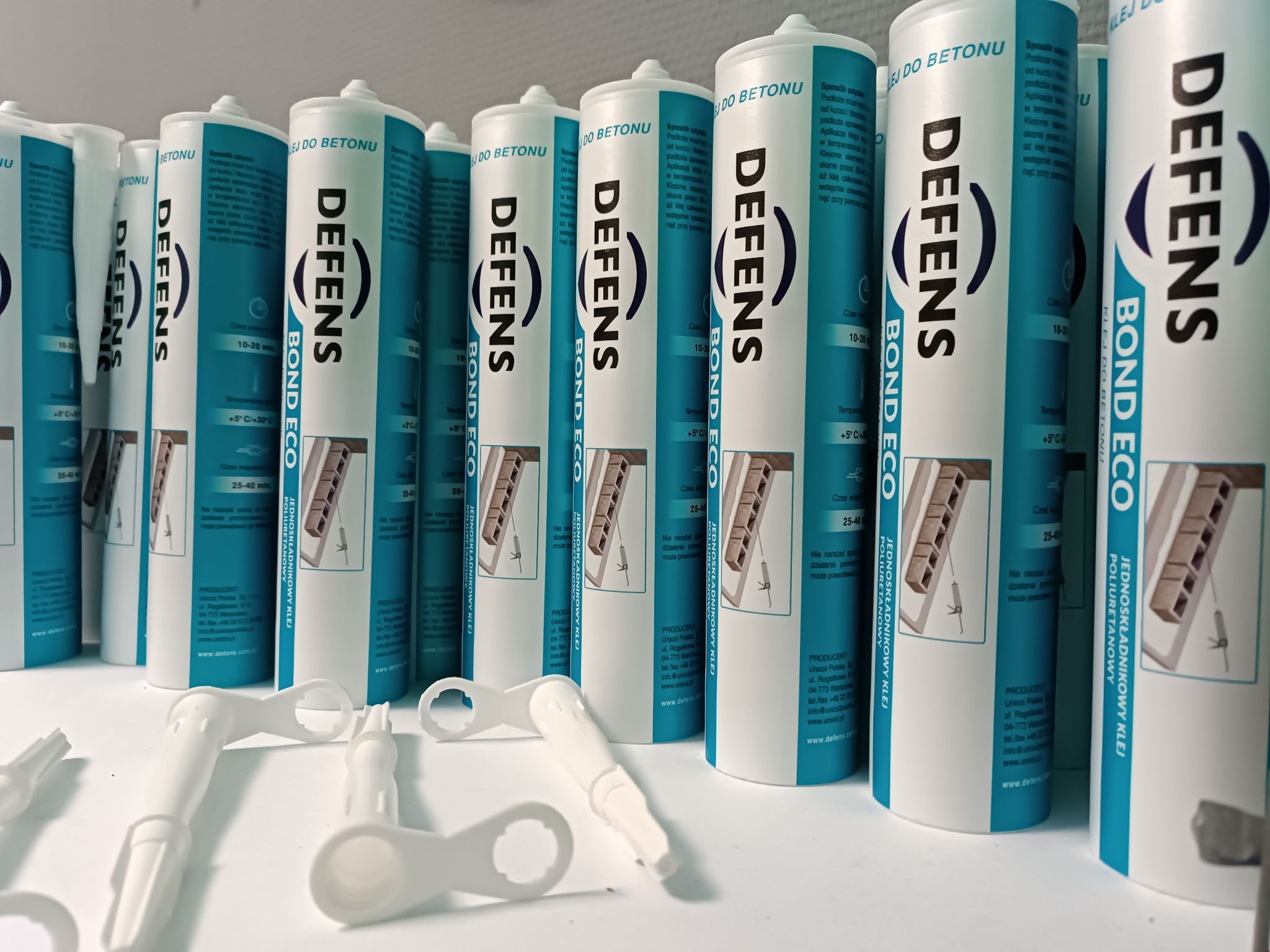 Rząd tub z klejem poliuretanowym do betonu marki DEFENS Bond Eco, z widocznymi aplikatorami, ustawione na białym blacie.