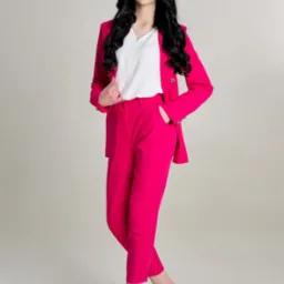 Portret kobiety w jaskraworóżowym garniturze i białych szpilkach na jasnym tle, pozującej z ręką w kieszeni spodni.