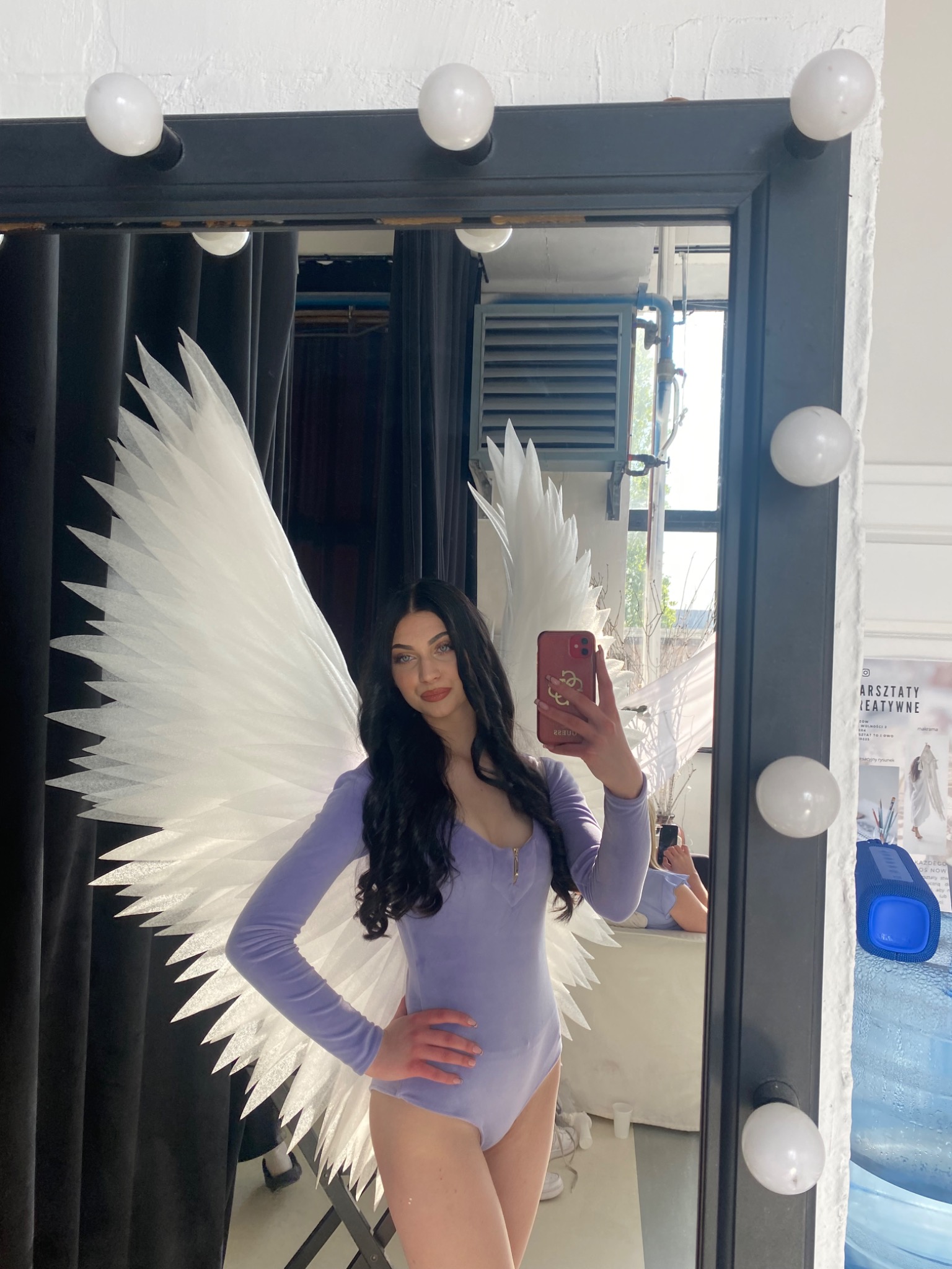 Kobieta w fioletowym body i białych skrzydłach anioła pozuje do selfie w lustrze z żarówkami, widoczny fragment garderoby i butelka wody.