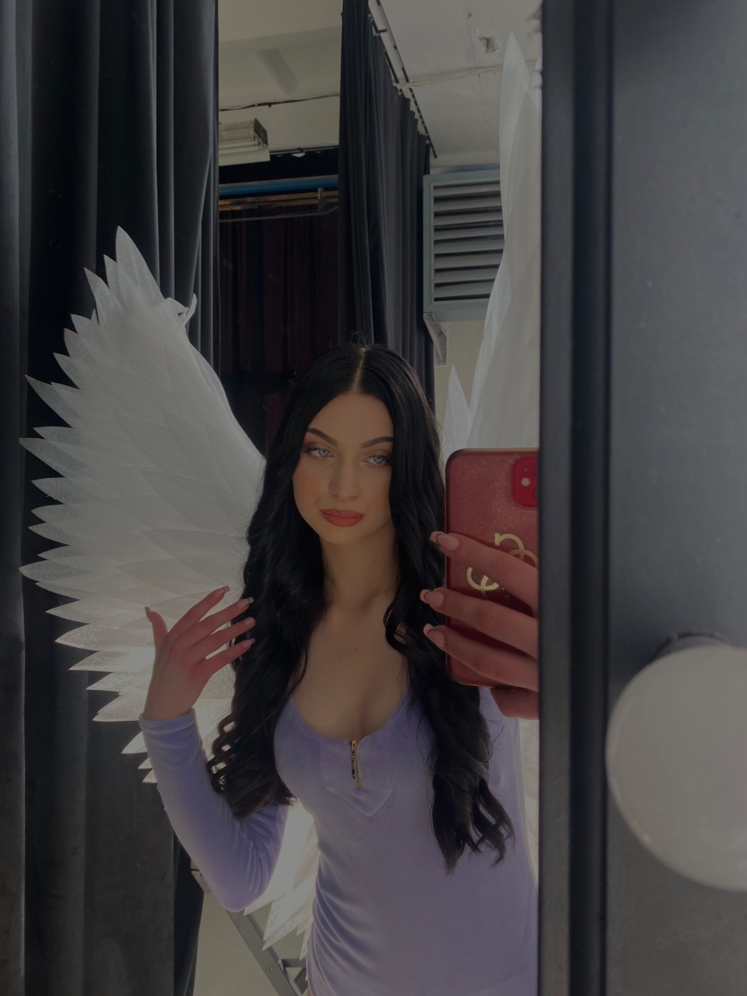 Portret kobiety z białymi skrzydłami anioła, robiącej selfie w lustrze garderoby z czarnymi zasłonami w tle.
