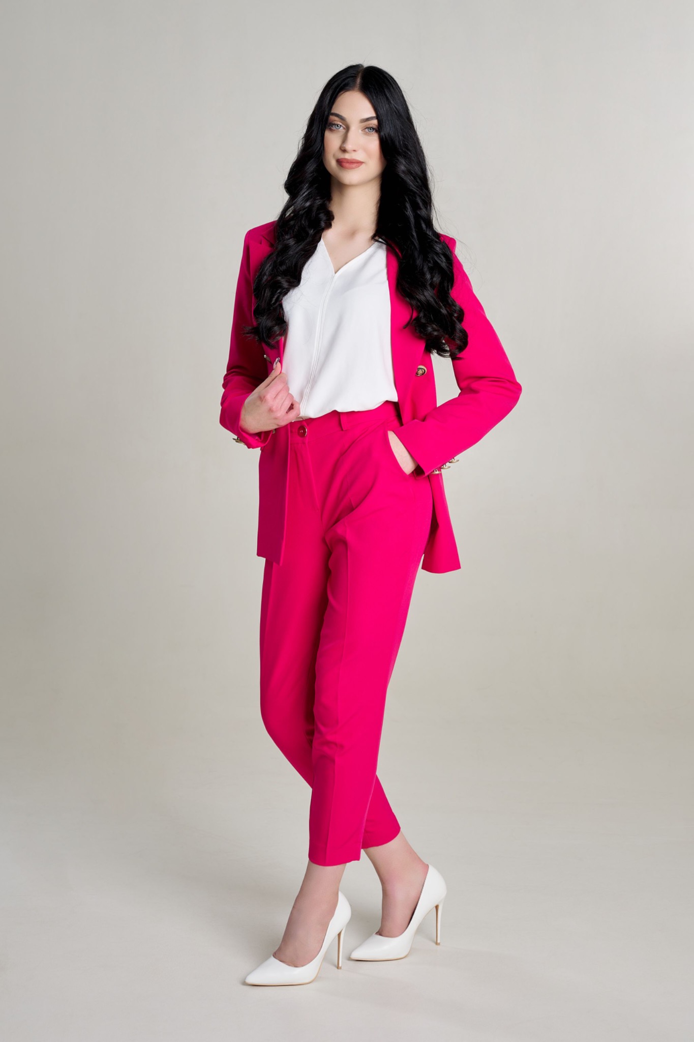 Portret kobiety w jaskraworóżowym garniturze i białych szpilkach na jasnym tle, pozującej z ręką w kieszeni spodni.