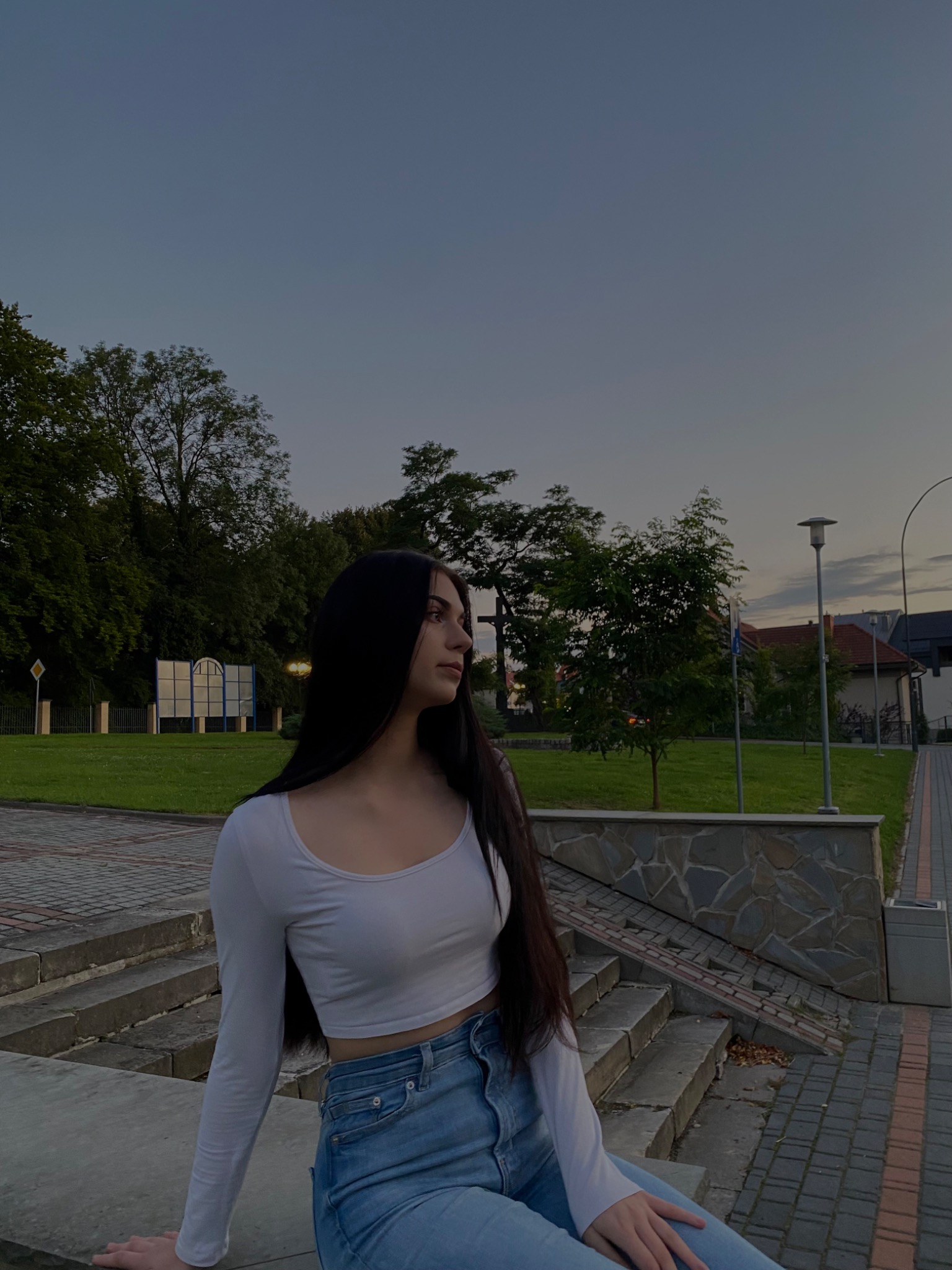 Portret młodej kobiety z długimi, ciemnymi włosami, siedzącej na betonowych schodach w parku o zmierzchu, ubrana w białą bluzkę z długim rękawem i dżinsy.