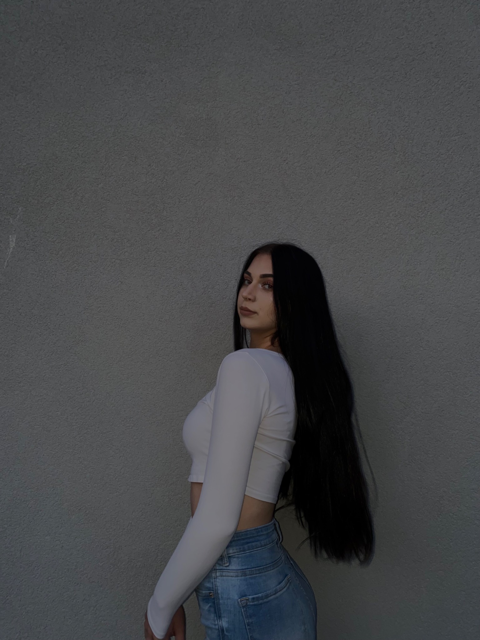 Portret kobiety z długimi, czarnymi włosami, odwróconej przez ramię w białym topie i dżinsach na tle szarej ściany.