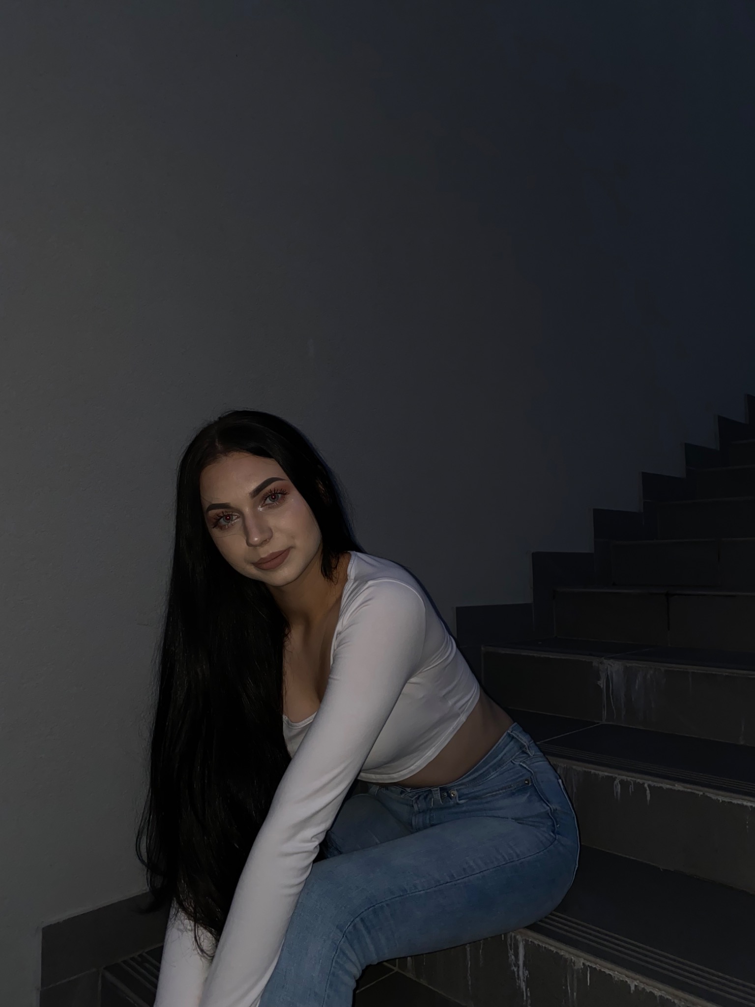 Młoda kobieta z długimi, czarnymi włosami, ubrana w białą bluzkę z długim rękawem i jasne dżinsy, siedzi na szarych schodach w słabym oświetleniu.