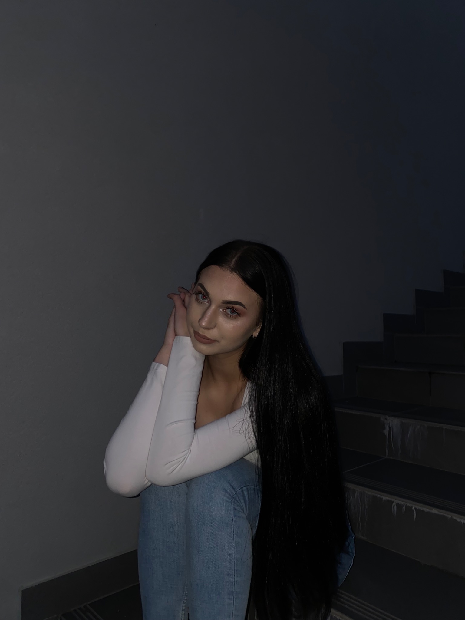 Portret młodej kobiety z długimi czarnymi włosami, w jasnej bluzce i dżinsach, siedzącej na schodach w ciemnym otoczeniu, z rękami podpartymi pod brodą.