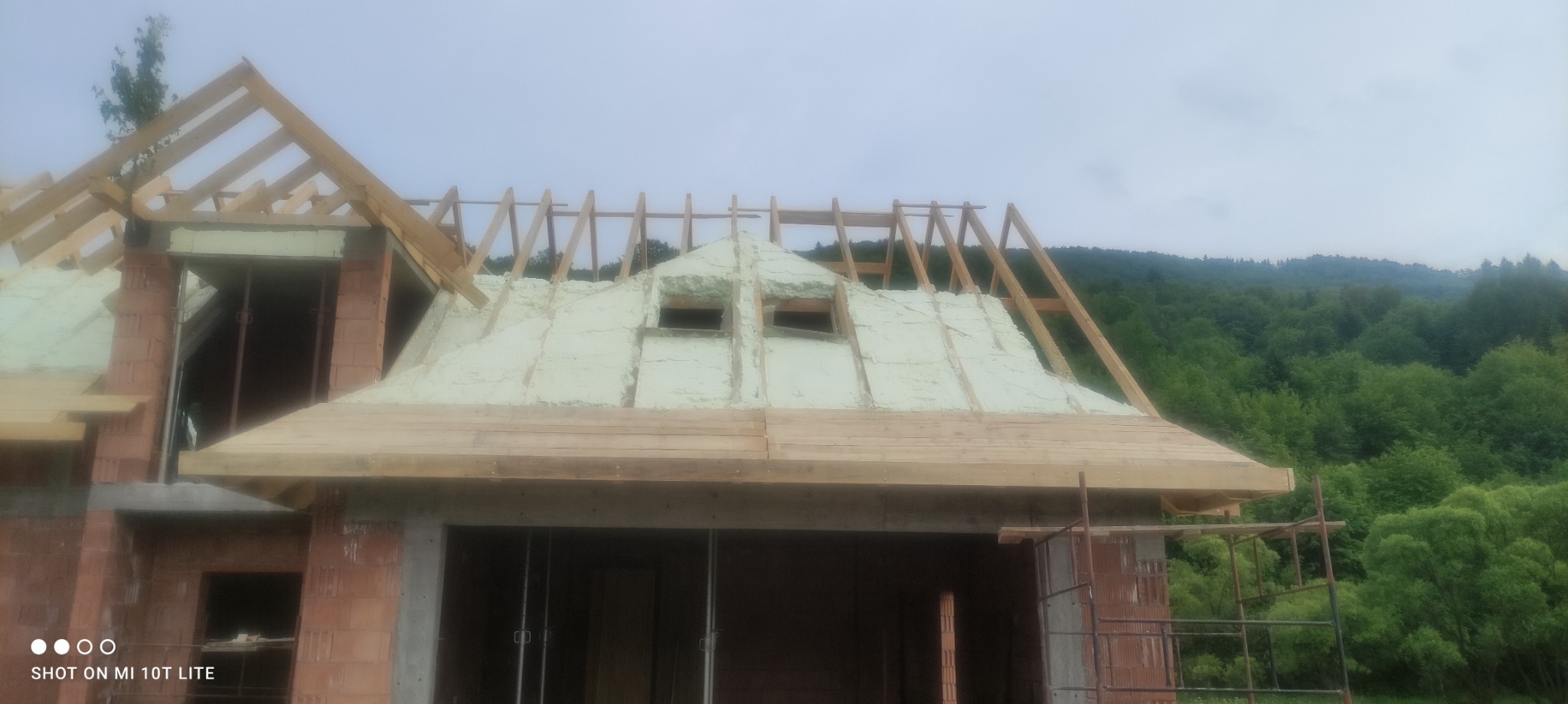 Niedokończony dom z drewnianą konstrukcją dachu i jasną, natryskową izolacją na poddaszu, widok na zalesione wzgórza w tle.