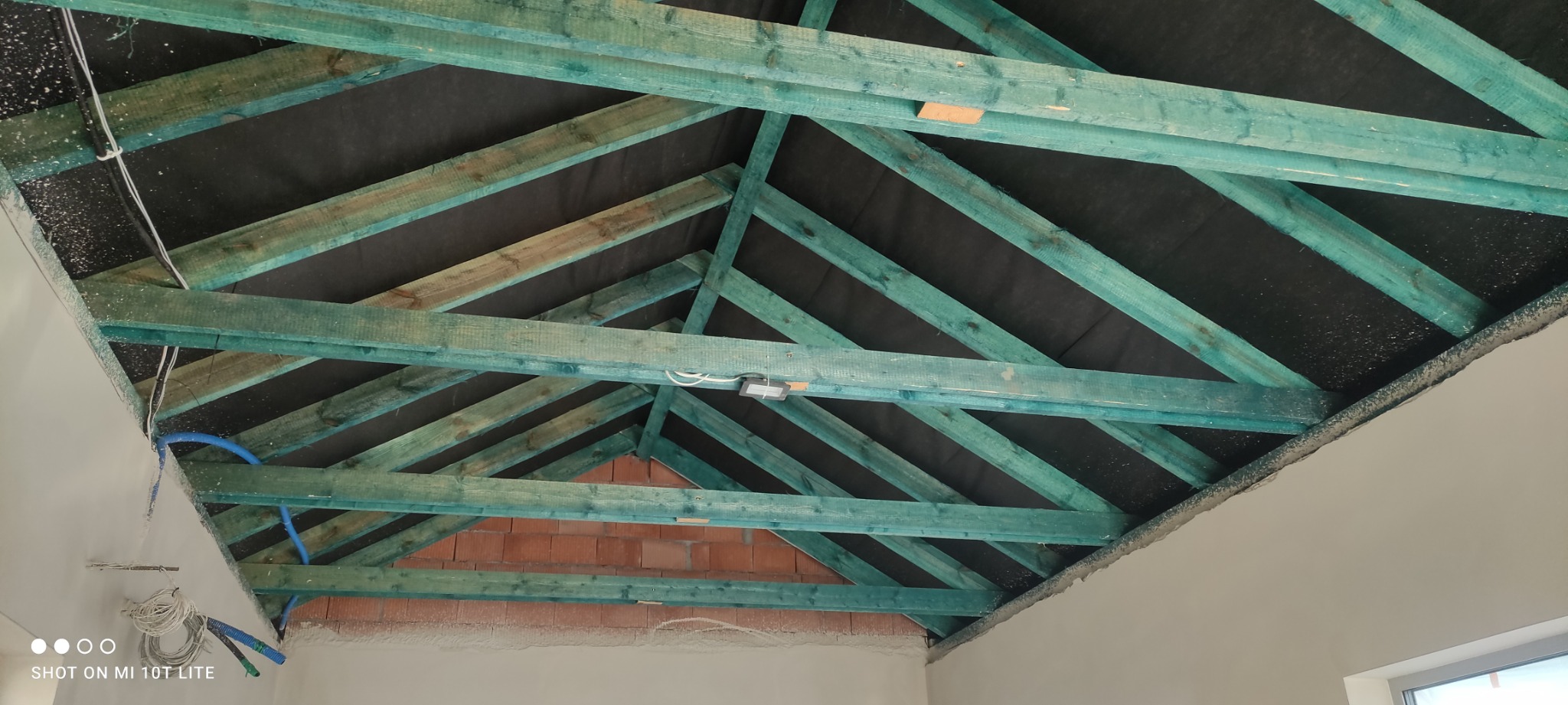 Skośny dach od wewnątrz z widoczną konstrukcją drewnianą pomalowaną na zielono-niebieski kolor, czarną membraną, instalacją elektryczną i fragmentem ściany z czerwonej cegły.