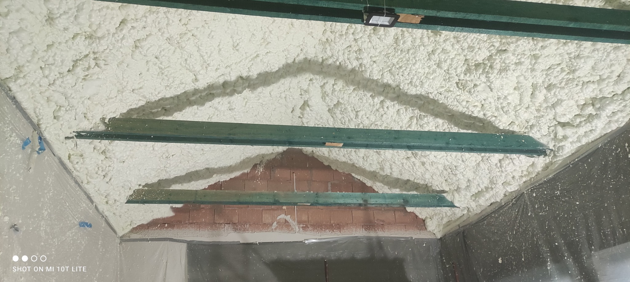 Poddasze z widoczną konstrukcją drewnianą i częściowo nałożoną białą pianką PUR izolacyjną, widok z dołu na sufit poddasza w trakcie ocieplania, z fragmentami odsłoniętej cegły.