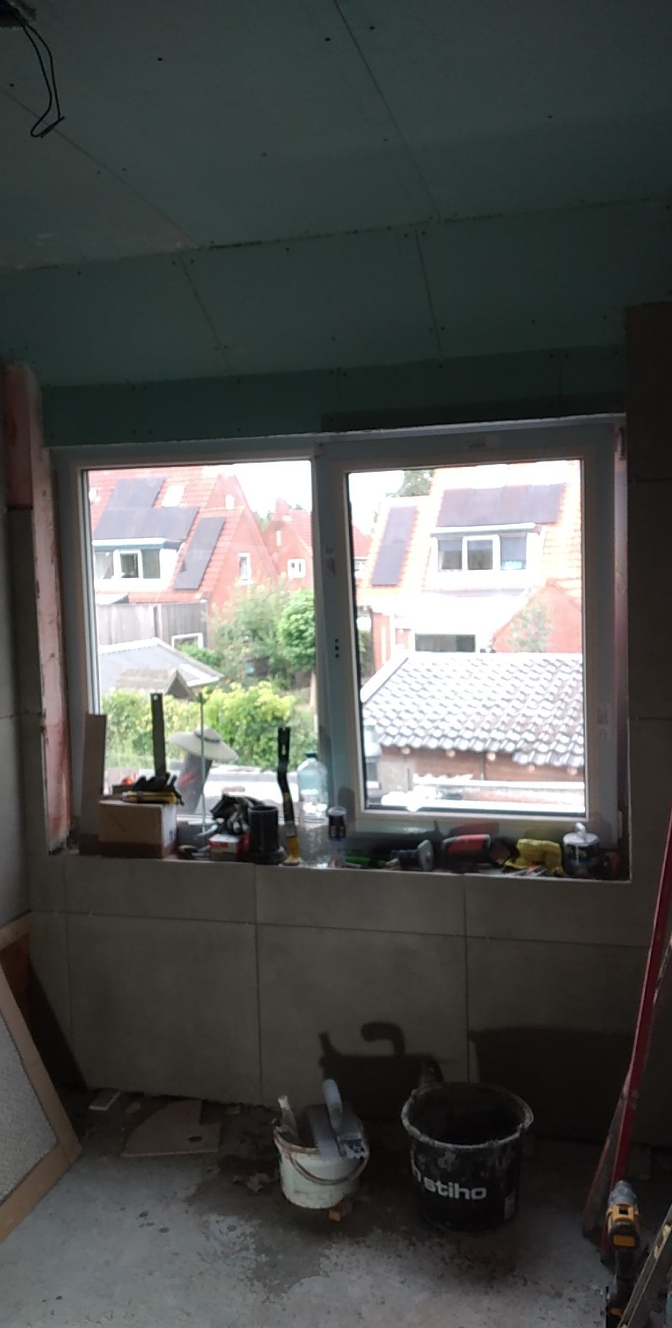 Pokój w trakcie prac wykończeniowych, widoczne płyty kartonowo-gipsowe na suficie, okno z widokiem na domy z panelami słonecznymi, narzędzia i wiadra na podłodze.