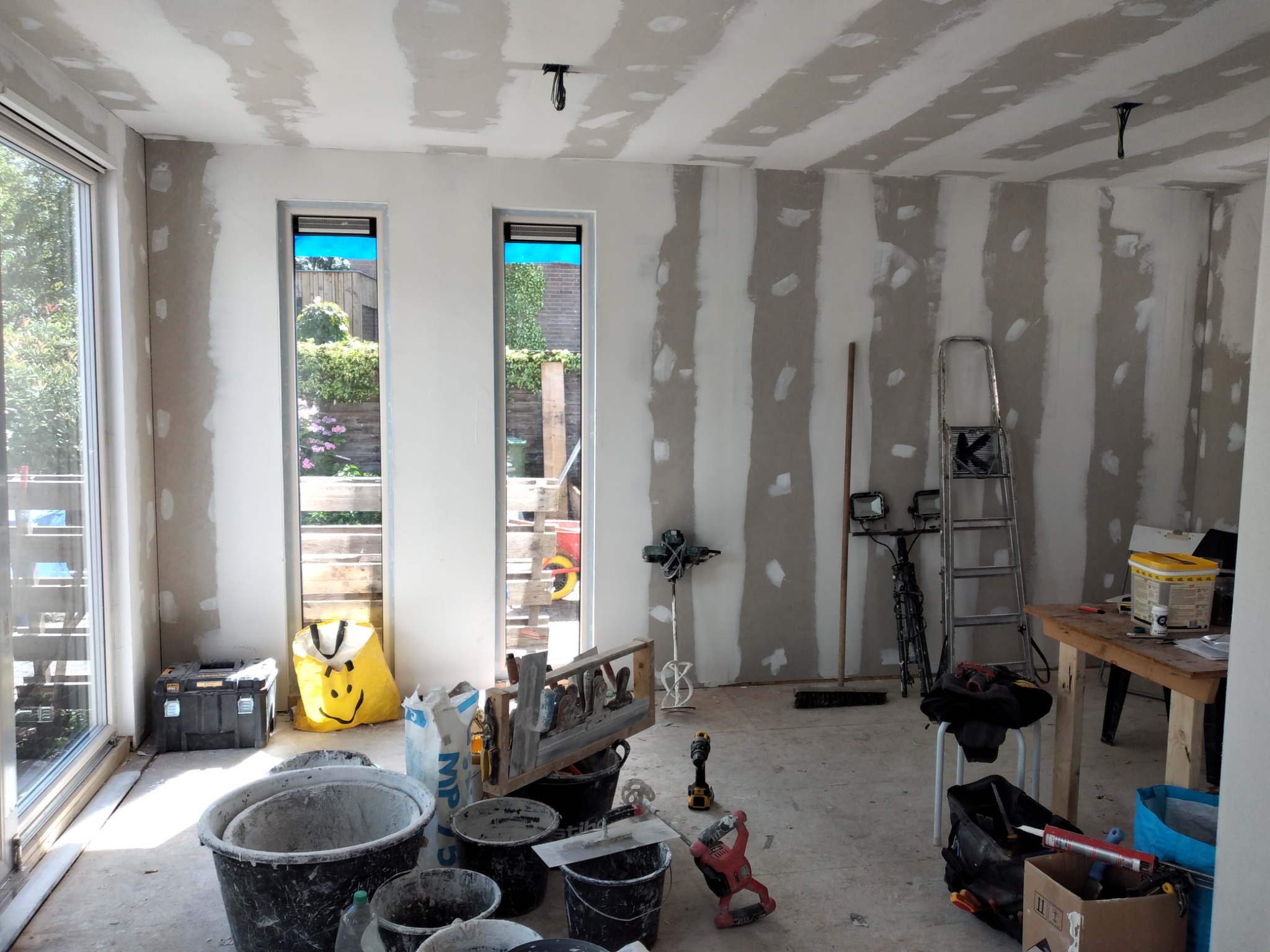 Wnętrze w trakcie prac wykończeniowych: ściany z płyt kartonowo-gipsowych z widocznymi łączeniami przygotowane do szpachlowania, liczne narzędzia budowlane, drabina, okna z widokiem na zieleń ogrodu.
