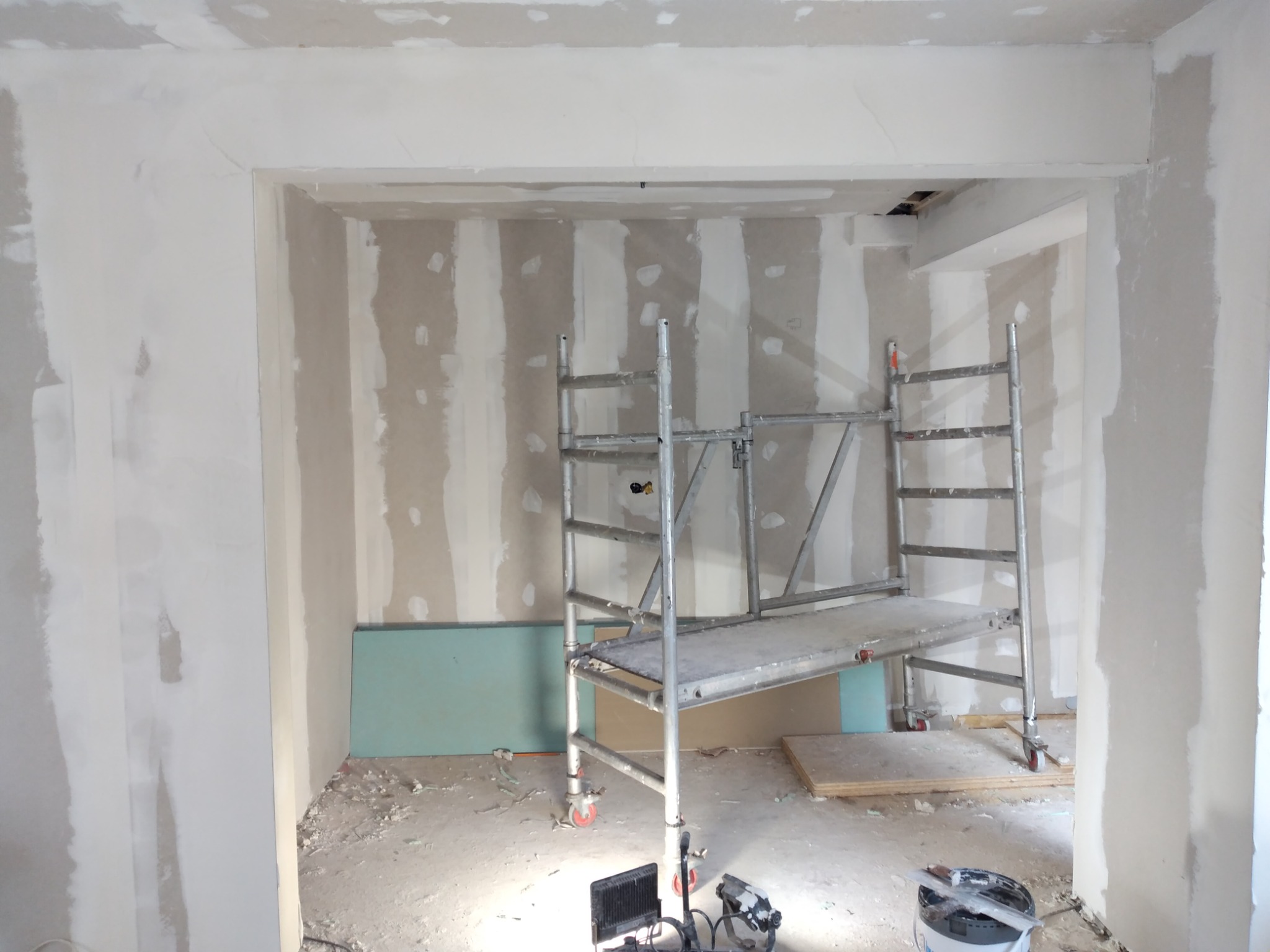 Wnętrze w trakcie prac przygotowawczych do malowania, widoczne ściany z nałożoną gładzią, częściowo surowe płyty kartonowo-gipsowe, metalowe rusztowanie, oświetlenie budowlane i wiadro z narzędziami.