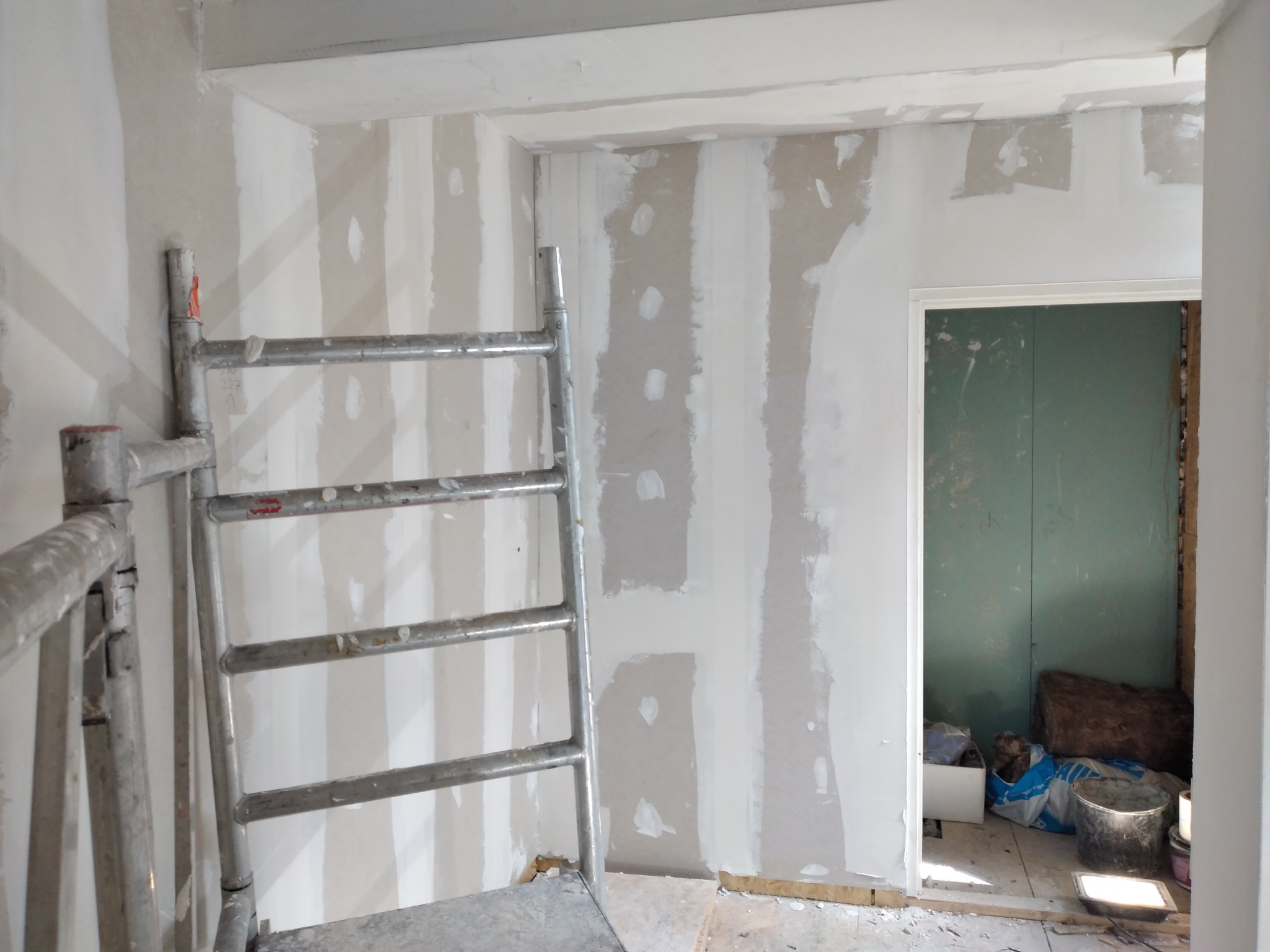 Metalowa drabina przystawiona do ściany w trakcie prac przygotowawczych, widoczne fragmenty płyt gipsowo-kartonowych i nałożona warstwa szpachli, w tle otwarte drzwi do innego pomieszczenia.