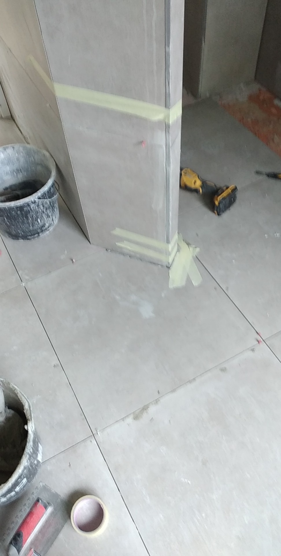 Układanie jasnych płytek podłogowych w trakcie prac wykończeniowych, widoczne narzędzia, wiadro z zaprawą i taśma malarska zabezpieczająca narożniki.