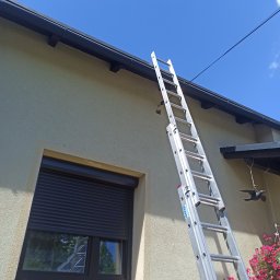 lublin.host KAROL RÓŻAŃSKI - Aluminiowa drabina oparta o ścianę domu z oknem i czarną rynną, błękitne niebo w tle. Widok z dołu, sugerujący prace na wysokościach.