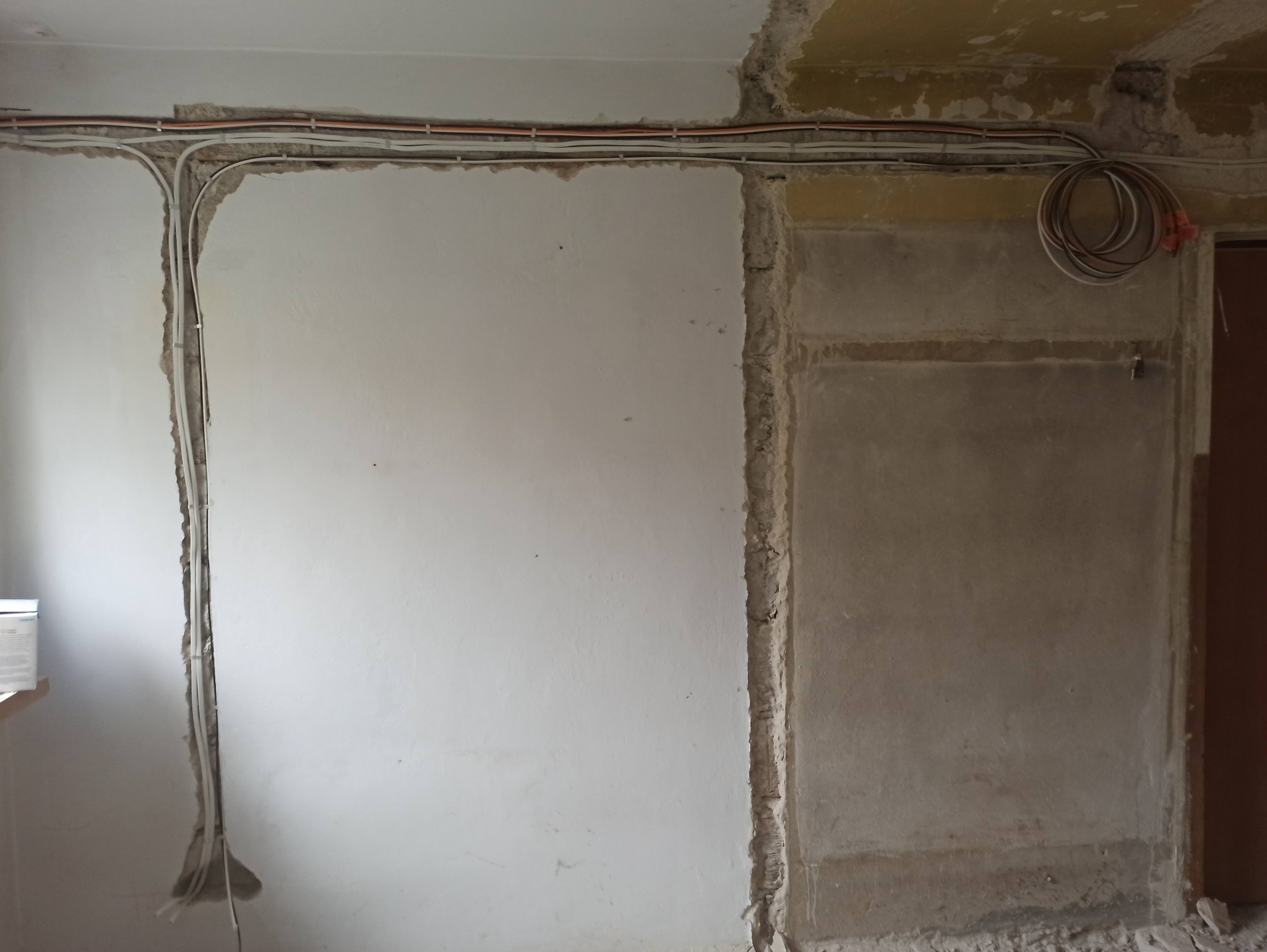 Instalacja elektryczna w trakcie remontu: kable prowadzone w bruzdach w ścianie, widoczne przewody pod sufitem i zwój kabli. Surowe wykończenie ścian.