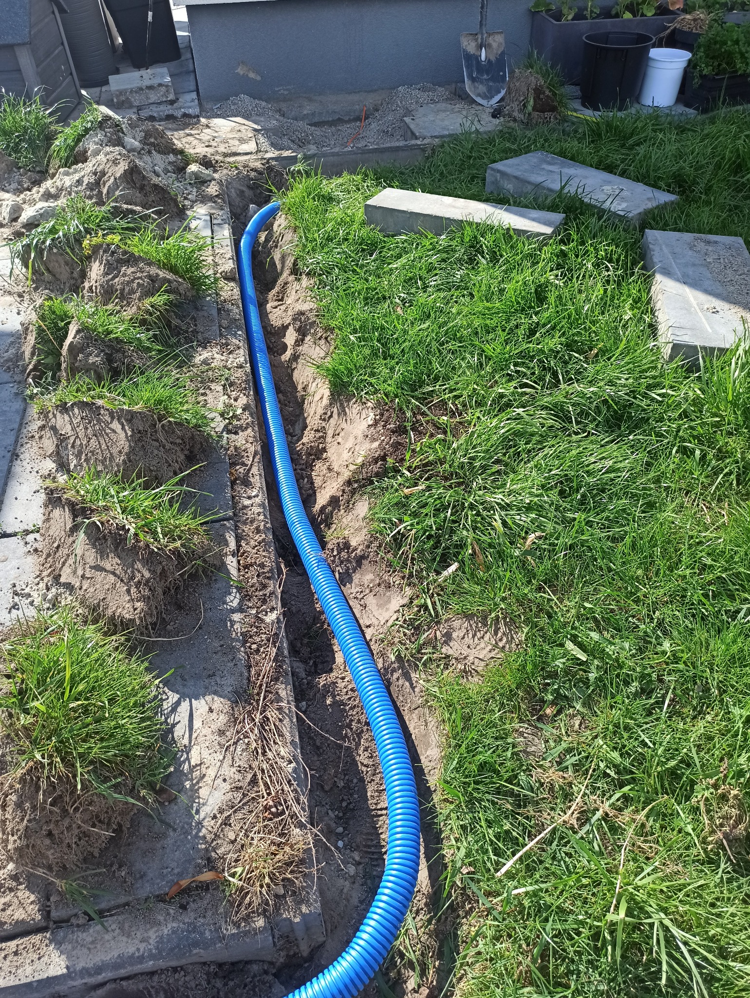 Niebieska rura karbowana ułożona w wykopie w trawie, prawdopodobnie instalacja elektryczna zewnętrzna lub drenaż. Widoczne narzędzia i materiały budowlane w tle.