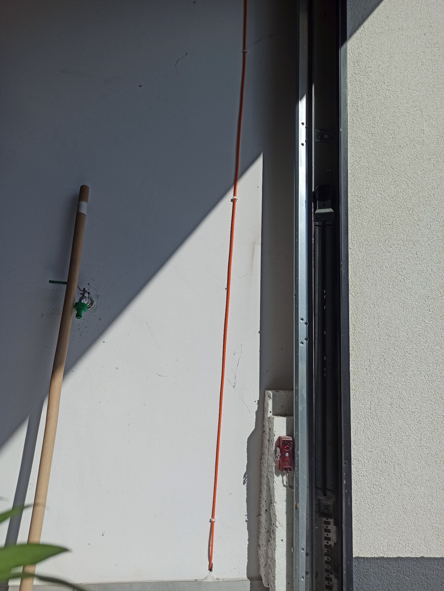 Pomarańczowy przewód przymocowany do białej ściany obok metalowej konstrukcji bramy garażowej z widocznymi sprężynami i czujnikiem. Ujęcie w dziennym świetle.