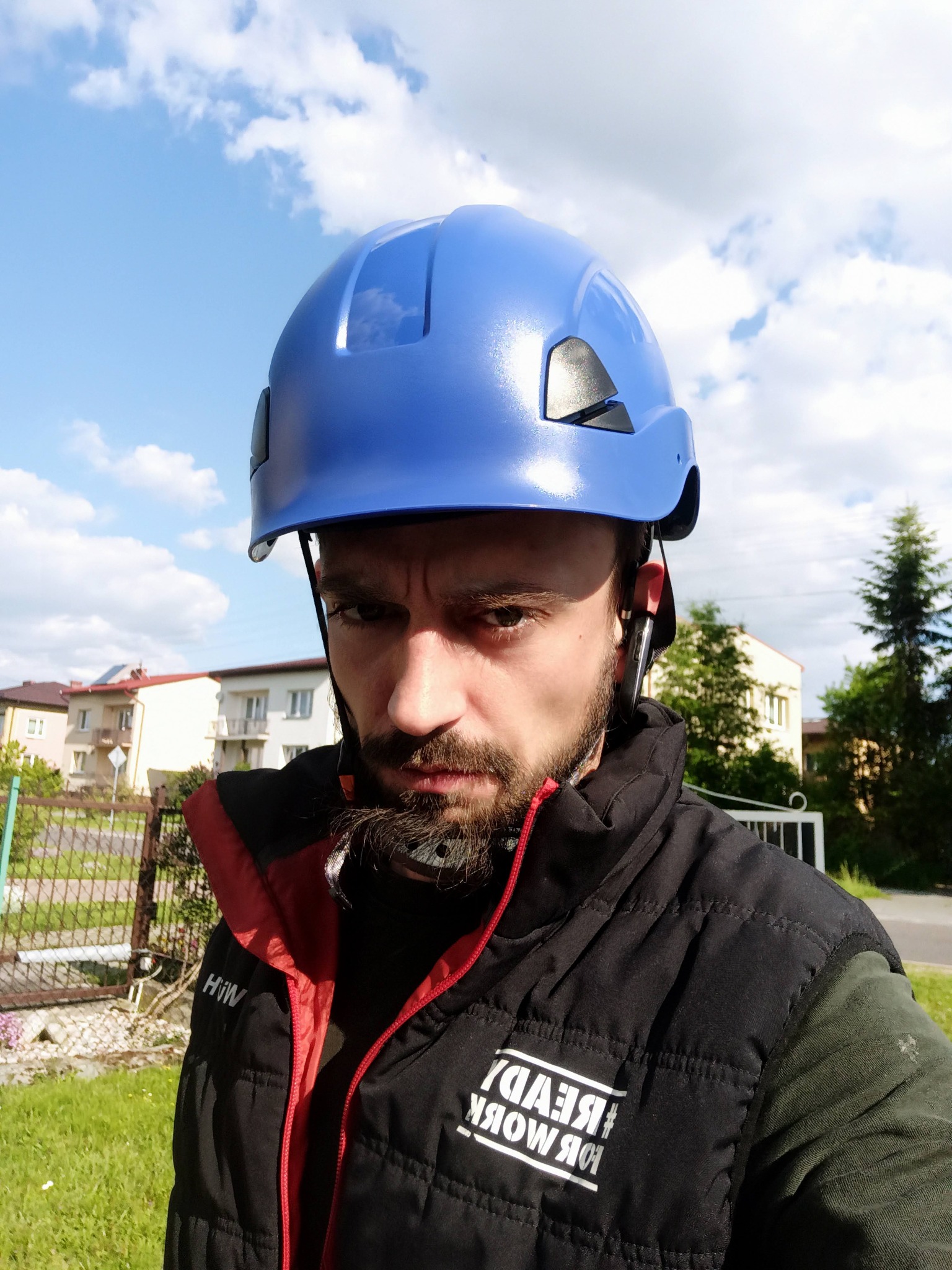 Mężczyzna w niebieskim kasku ochronnym, czarnej kamizelce z napisem #READY FOR WORK, na tle błękitnego nieba i zabudowań. Ujęcie z dołu.