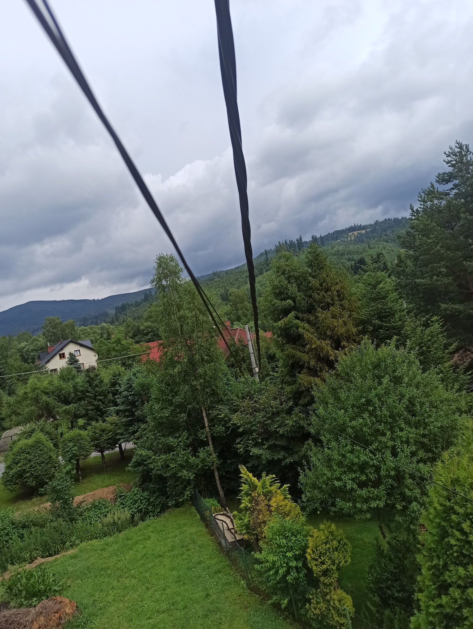 Widok z góry na zalesione wzgórza i dom z przewodami elektrycznymi na pierwszym planie, pochmurne niebo w tle. Ujęcie z perspektywy instalacji elektrycznej.