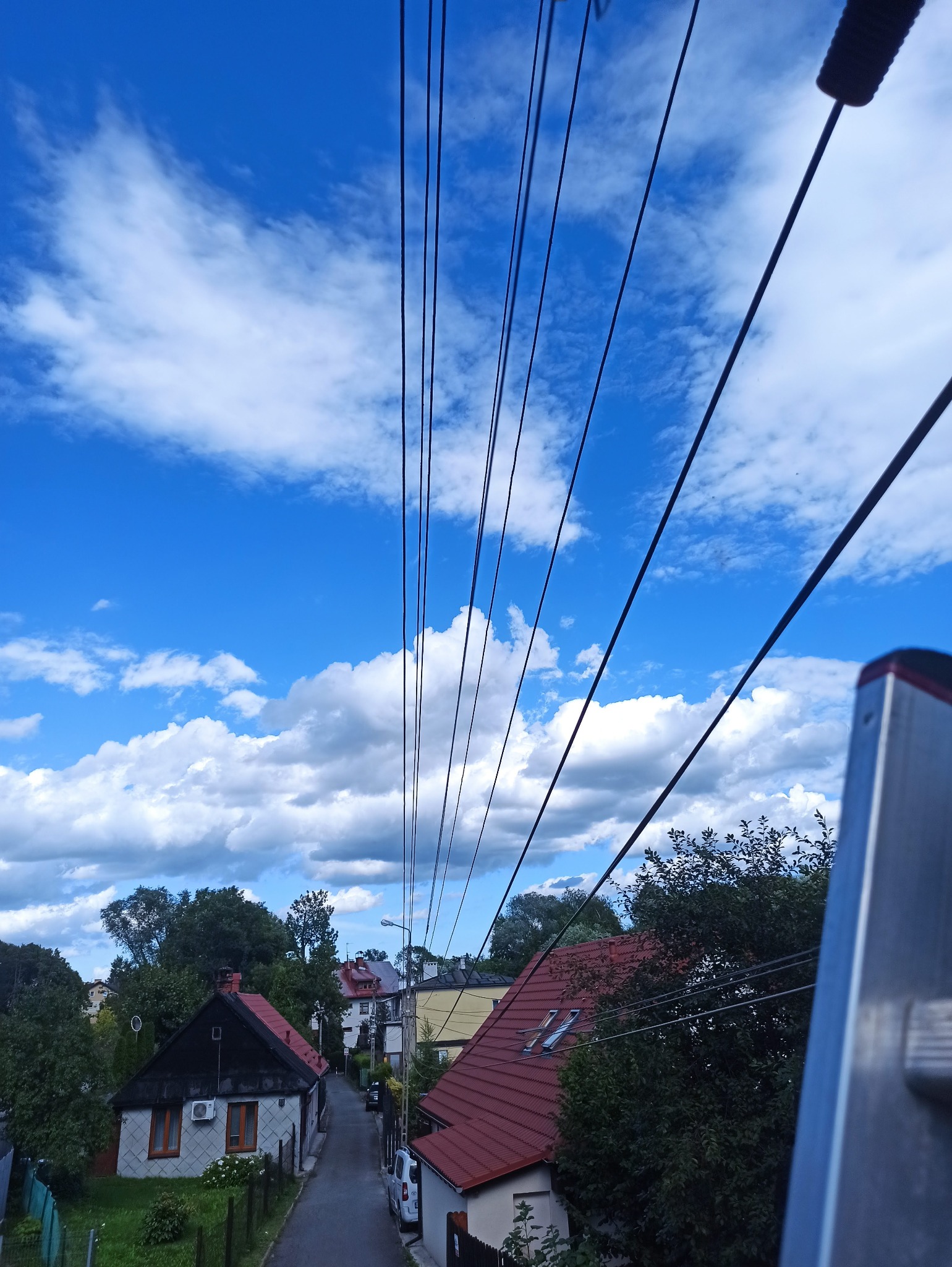 Słupy i przewody elektryczne na tle błękitnego nieba z chmurami, widoczne z perspektywy drabiny. Ujęcie z dołu, w tle zabudowa jednorodzinna.