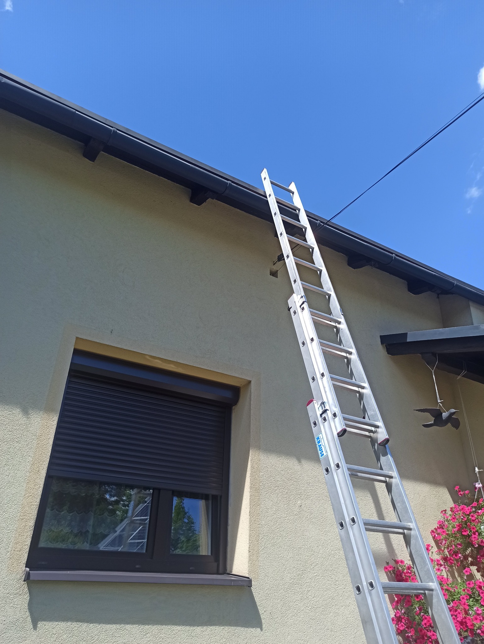 Aluminiowa drabina oparta o ścianę domu z oknem i czarną rynną, błękitne niebo w tle. Widok z dołu, sugerujący prace na wysokościach.