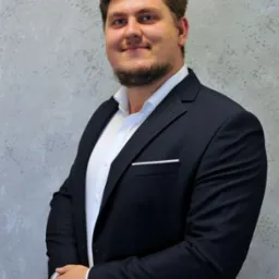 Portret uśmiechniętego mężczyzny w ciemnym garniturze i białej koszuli, z rękami skrzyżowanymi na wysokości talii, na tle szarej, teksturowanej ściany.