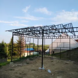 Szkielet metalowej konstrukcji w trakcie budowy, prawdopodobnie wiaty lub zadaszenia, na tle wiejskiego krajobrazu z domami i drzewami.