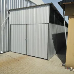 Metalowa brama wjazdowa z dwuskrzydłowymi drzwiami, wykonana z blachy falistej, z widocznymi śladami spawania i prostym zamkiem, na tle kostki brukowej i fragmentów innych budynków.