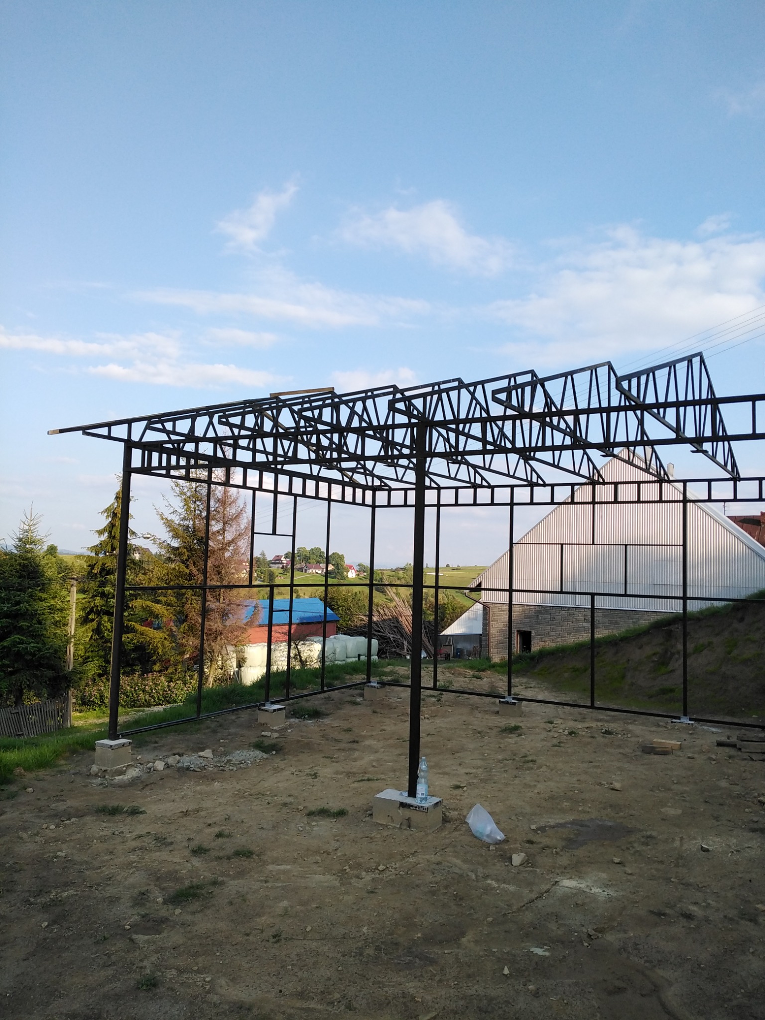 Szkielet metalowej konstrukcji w trakcie budowy, prawdopodobnie wiaty lub zadaszenia, na tle wiejskiego krajobrazu z domami i drzewami.