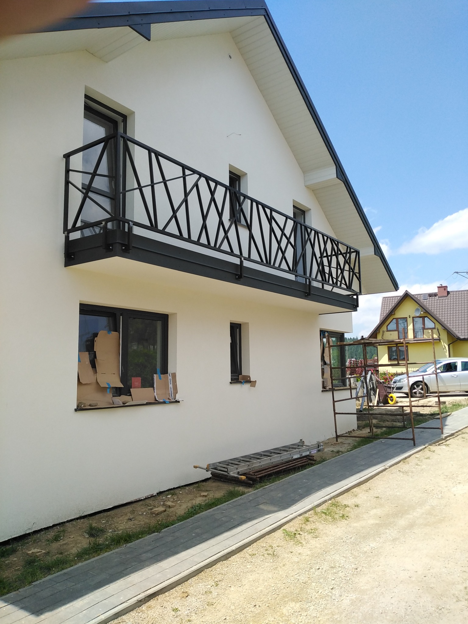 Biały dom z czarnym, geometrycznym balkonem z metalu, widoczne rusztowanie i drabina przy ścianie, okna zabezpieczone kartonem.