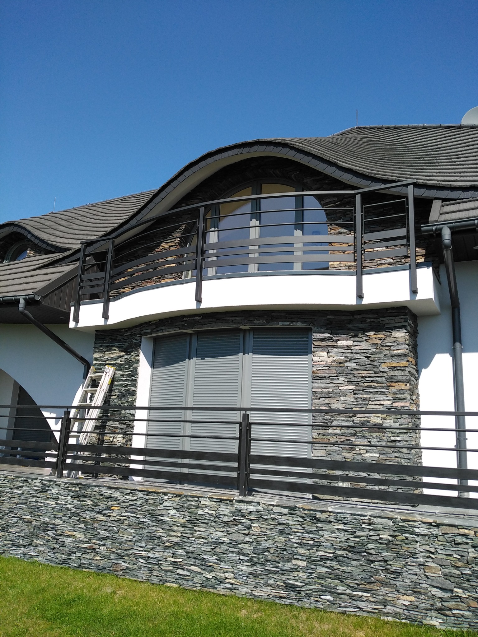 Nowoczesny dom z balkonem zabezpieczonym czarną, metalową balustradą i elewacją z kamienia łupkowego, z drabiną opartą o ścianę.