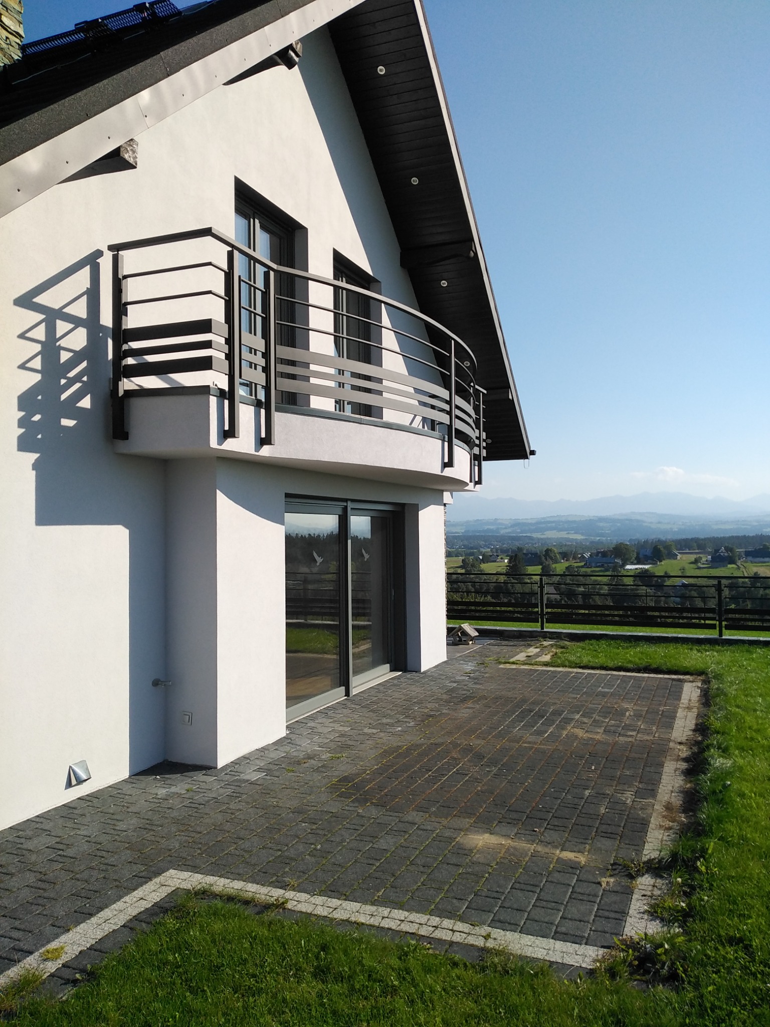 Nowoczesny dom z balkonem z metalową balustradą i tarasem wyłożonym kostką brukową, w tle widok na góry.