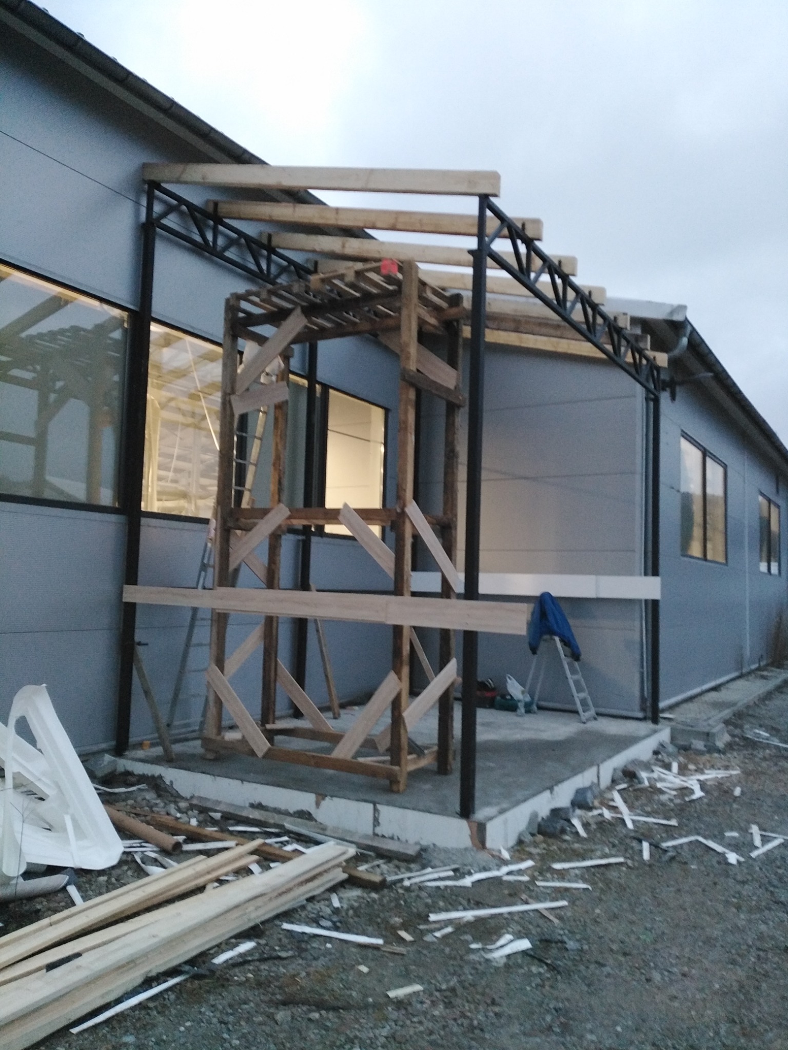 Montaż zadaszenia nad wejściem do budynku z szarej blachy trapezowej, widoczne elementy drewniane i metalowe konstrukcji, drabina oparta o ścianę, teren wokół budowy z resztkami materiałów.