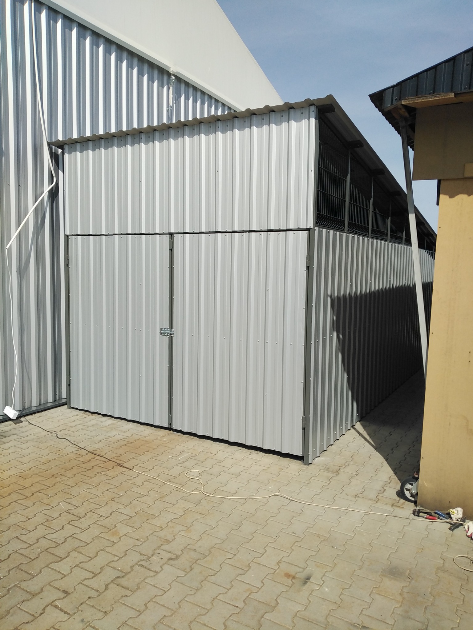 Metalowa brama wjazdowa z dwuskrzydłowymi drzwiami, wykonana z blachy falistej, z widocznymi śladami spawania i prostym zamkiem, na tle kostki brukowej i fragmentów innych budynków.