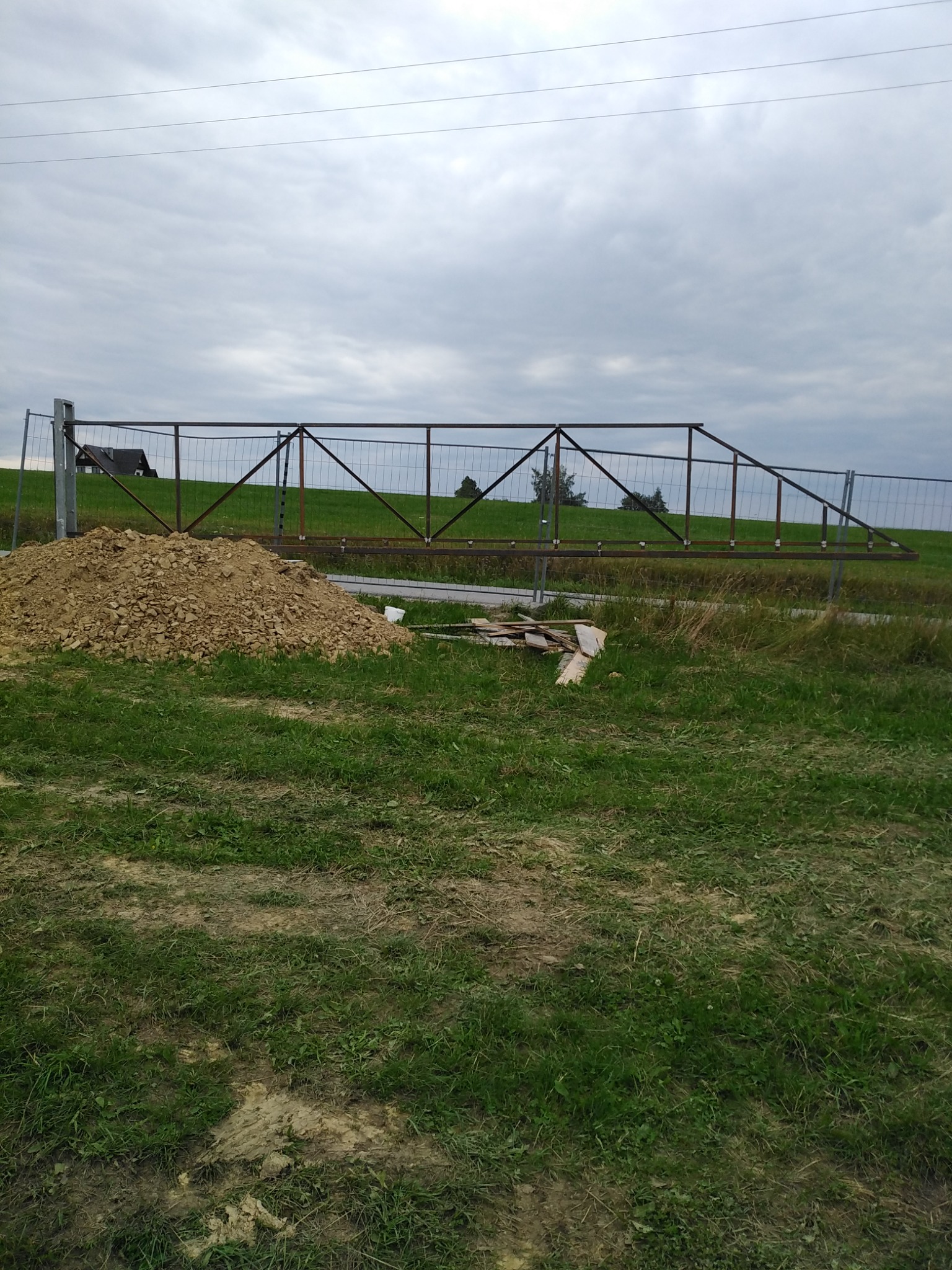 Surowa konstrukcja metalowej bramy wjazdowej w trakcie montażu, widoczny stos ziemi i tymczasowe ogrodzenie placu budowy na tle pochmurnego nieba.