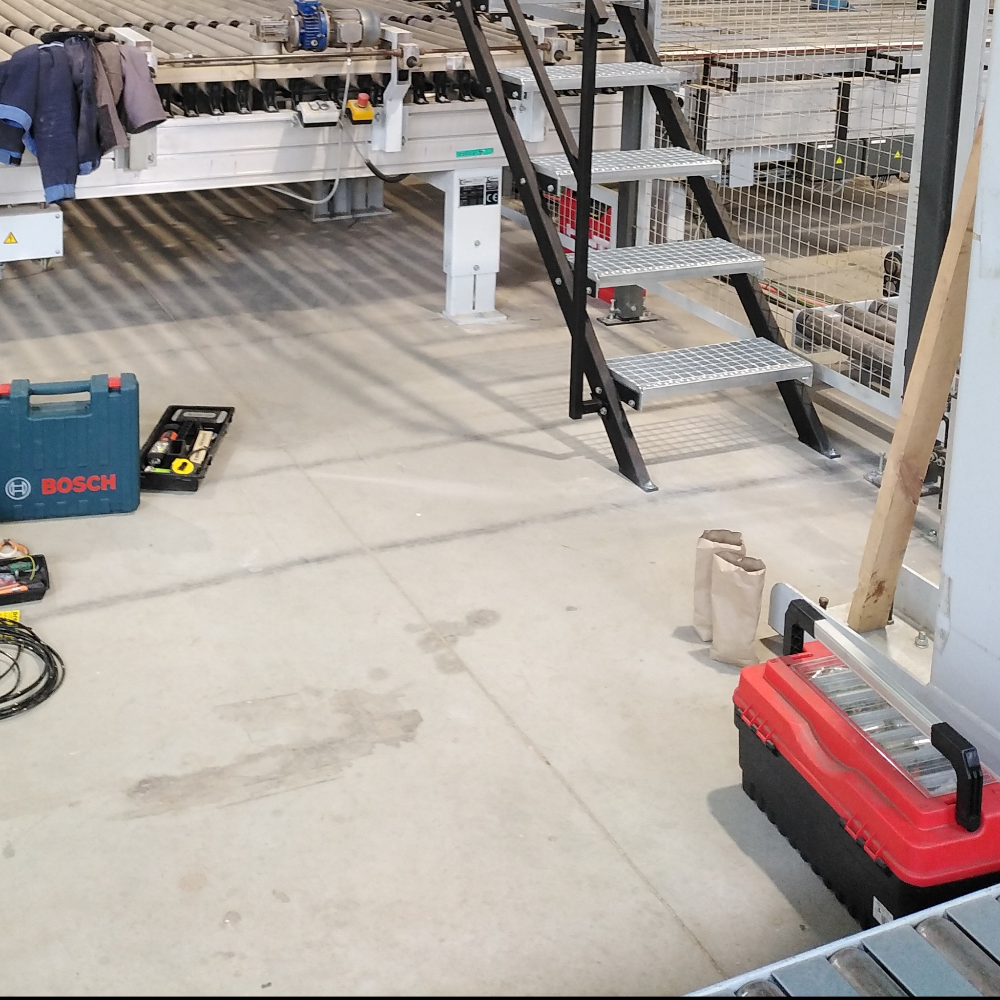 Widok hali produkcyjnej z metalową konstrukcją, schodami z kraty, skrzynkami narzędziowymi Bosch i czerwono-czarną skrzynką na narzędzia oraz elementami linii produkcyjnej.