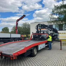 Dwaj mężczyźni w kamizelkach odblaskowych przy ciężarówce z platformą i dźwigiem HAP T 10-3, drugi dźwig MAN w tle, kostka brukowa na placu.