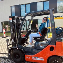 Centrum Szkoleniowe LDM w Legnicy - Uśmiechnięta kobieta w okularach przeciwsłonecznych siedzi za kierownicą pomarańczowo-szarego wózka widłowego Toyota Tonero 15 na kostce brukowej, w tle budynek z ciemnymi oknami i błękitne niebo.