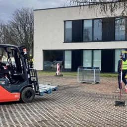 Grupa osób w kamizelkach odblaskowych obserwuje szkolenie z obsługi wózka widłowego Toyota Tonero 15 na placu manewrowym, w tle podnośnik koszowy i budynek.