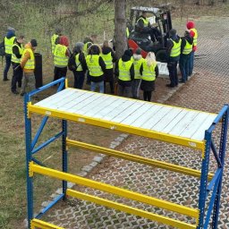 Centrum Szkoleniowe LDM w Legnicy - Grupa osób w kamizelkach odblaskowych obserwuje pracę wózka widłowego obok regału magazynowego z białymi półkami.