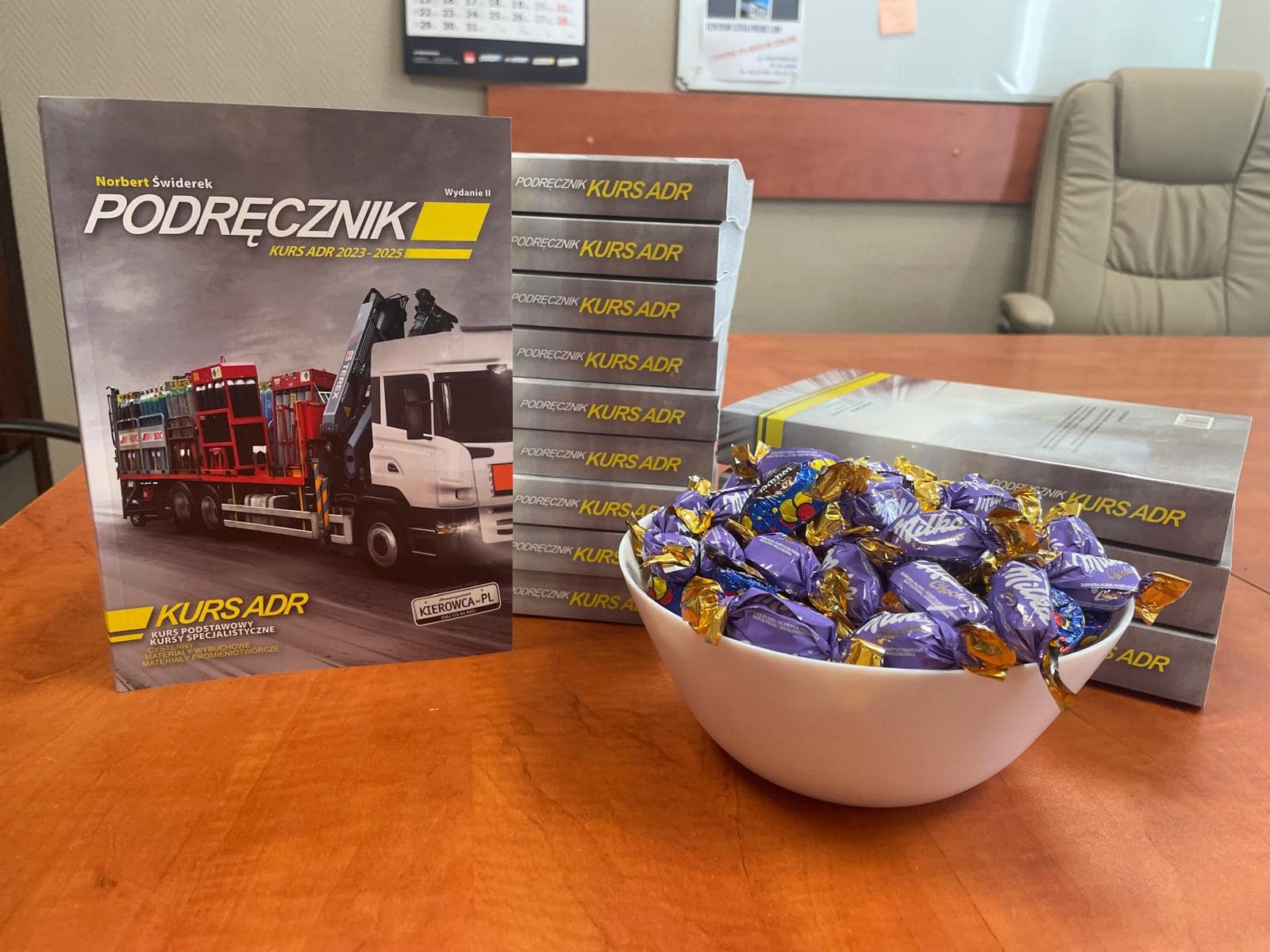 Podręcznik 'Kurs ADR' autorstwa Norberta Świderka, wydanie II, obok stos książek i miska pełna cukierków Milka na drewnianym biurku.