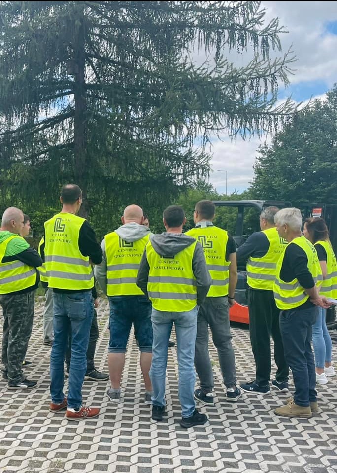 Grupa osób w kamizelkach odblaskowych z napisem 'Centrum Szkoleniowe' stoi na zewnątrz, prawdopodobnie przed wózkiem widłowym, otoczona zielenią.