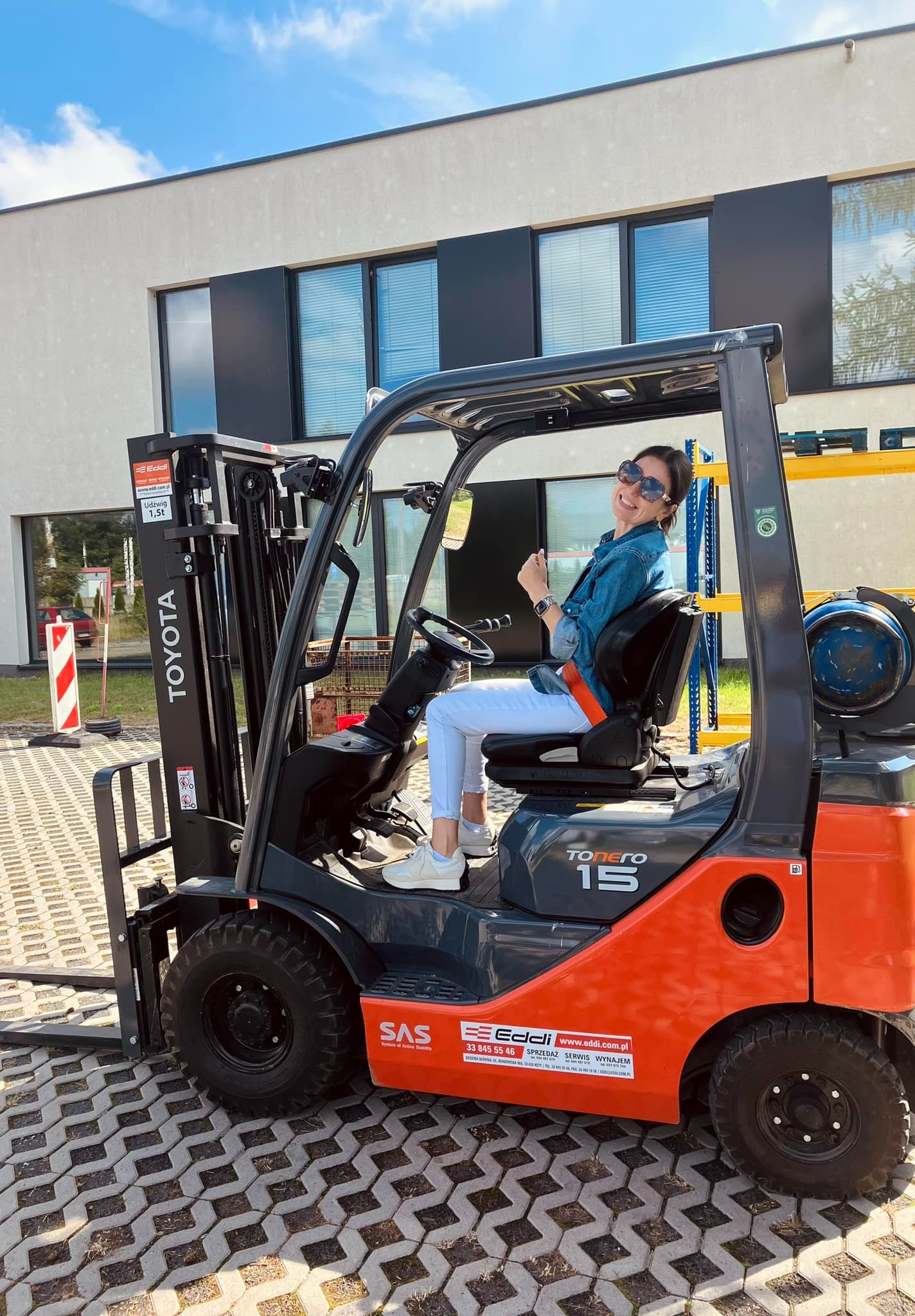Uśmiechnięta kobieta w okularach przeciwsłonecznych siedzi za kierownicą pomarańczowo-szarego wózka widłowego Toyota Tonero 15 na kostce brukowej, w tle budynek z ciemnymi oknami i błękitne niebo.