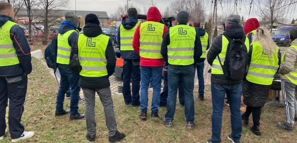 Grupa osób w kamizelkach odblaskowych z logo 'LDM Centrum Szkoleniowe' stoi na zewnątrz, prawdopodobnie przed wózkiem widłowym, podczas szkolenia.