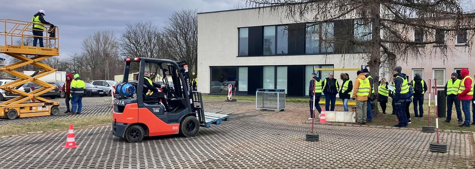 Grupa osób w kamizelkach odblaskowych obserwuje szkolenie z obsługi wózka widłowego Toyota Tonero 15 na placu manewrowym, w tle podnośnik koszowy i budynek.