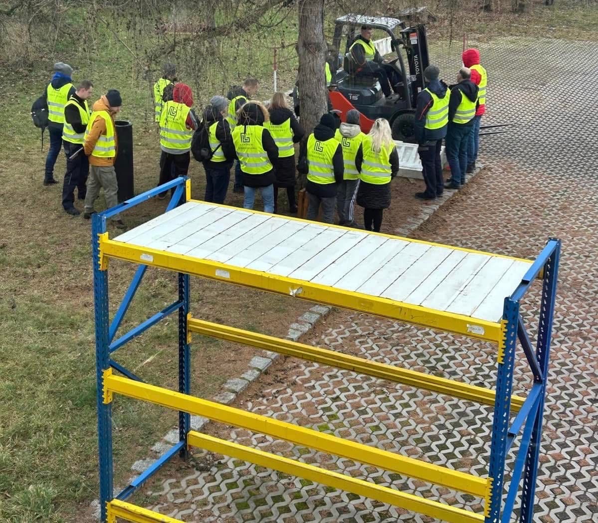 Grupa osób w kamizelkach odblaskowych obserwuje pracę wózka widłowego obok regału magazynowego z białymi półkami.