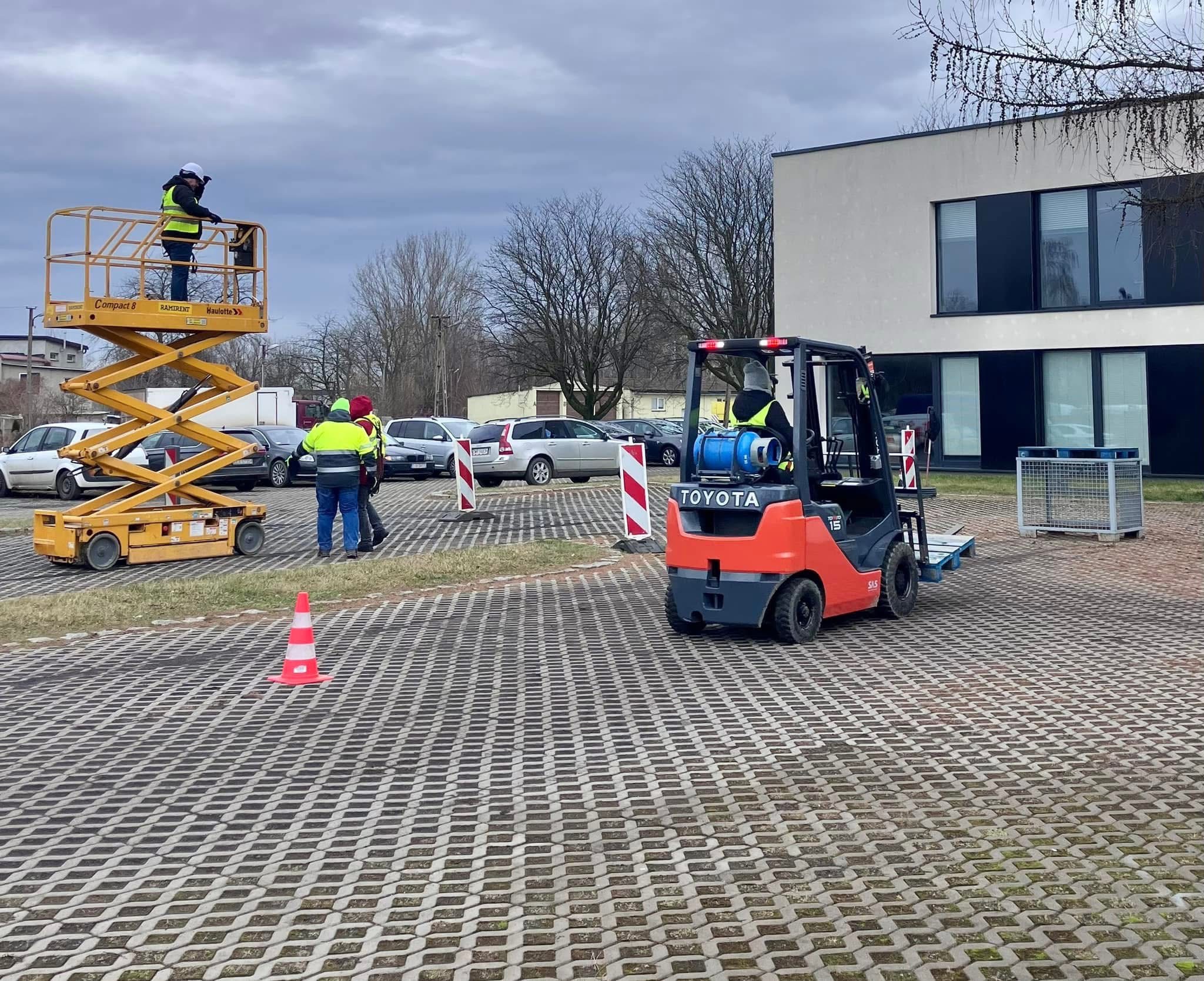 Pracownik na podnośniku koszowym Compact 8 i operator wózka widłowego Toyota na placu budowy, obok stoją zaparkowane samochody i budynek z dużymi oknami.